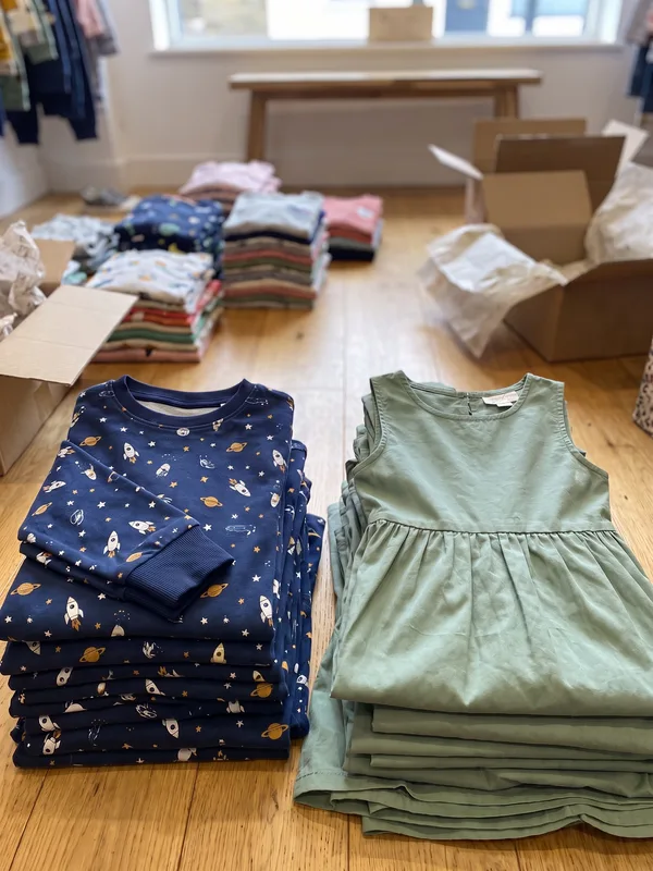 Pilhas de roupa infantil recém-chegada no piso da loja, com um pijama estampado de foguetes e um vestido verde no topo